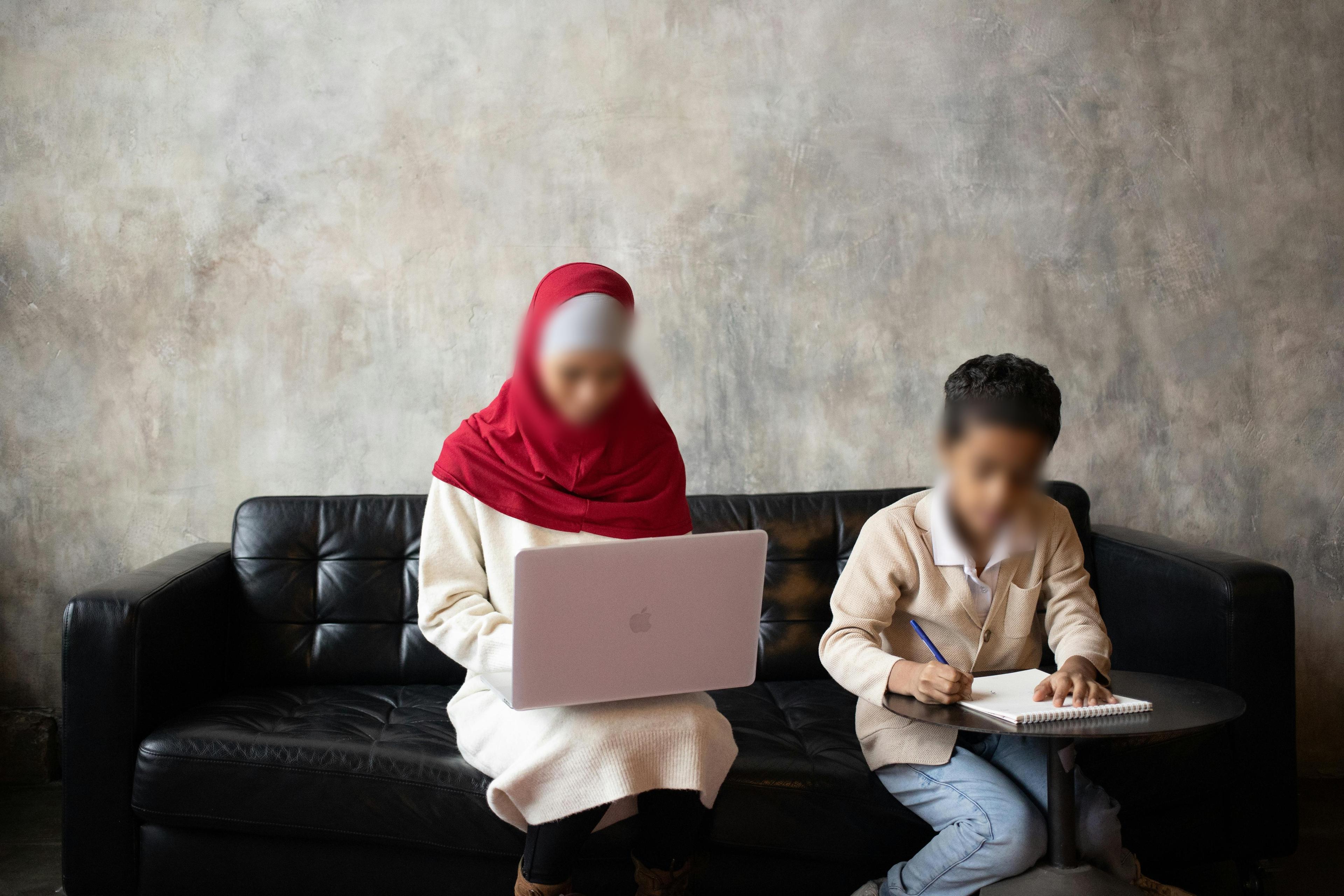 A person wearing a red hijab uses a laptop on a black sofa, while a child sits beside them writing in a notebook at a small table. Their faces are blurred. The background is a plain, light-colored wall.
