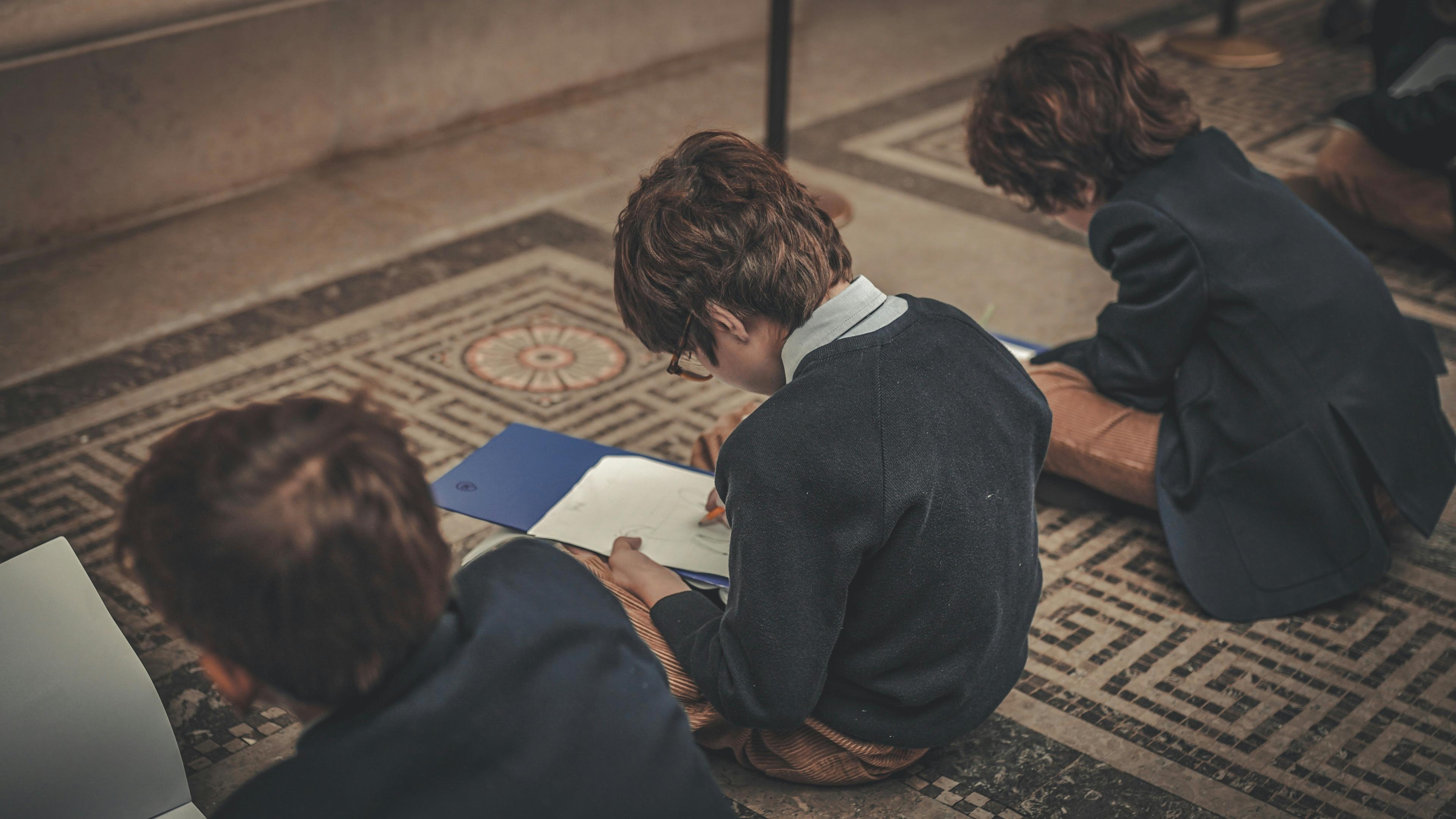Three children sit on a tiled floor, each wearing dark jackets and brown pants, focused on writing or drawing on white papers placed on blue folders. Their backs are turned to the camera.