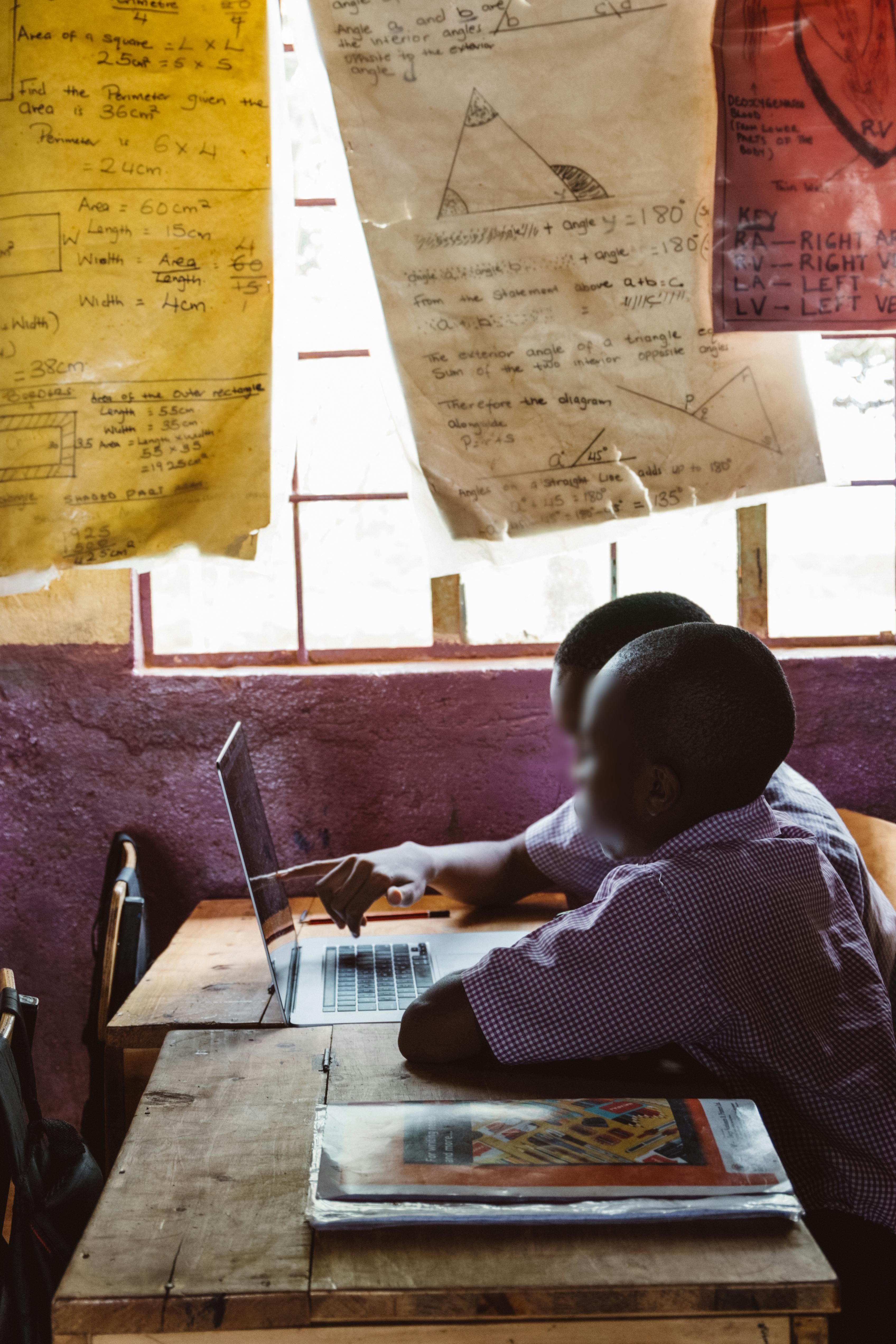 Two students in a classroom focus on a laptop. Charts with geometric figures hang above them. The atmosphere conveys learning and collaboration.