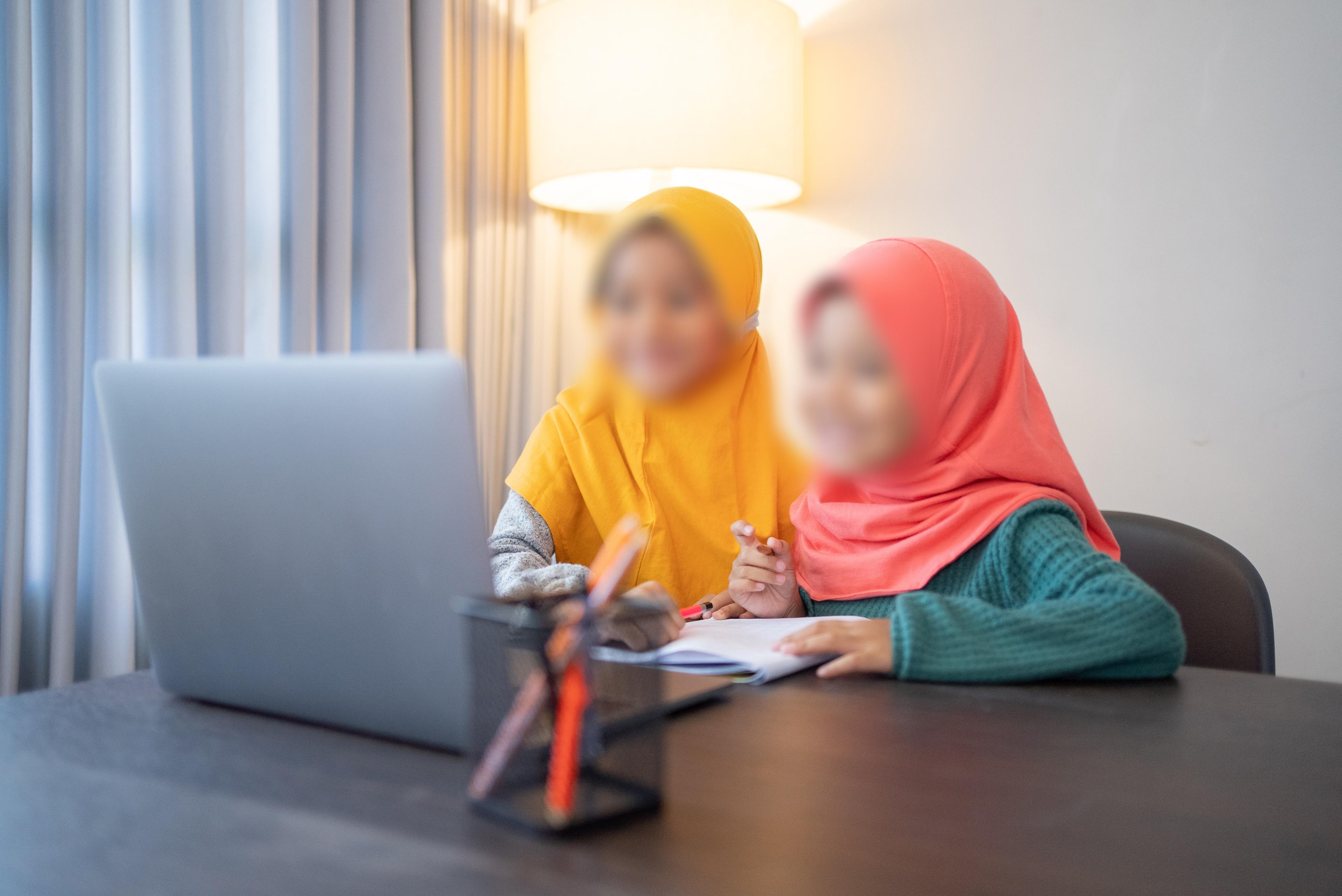Two children wearing hijabs sit at a desk, working together on schoolwork with a laptop and notebook. A lamp glows in the background, creating a cozy study environment.